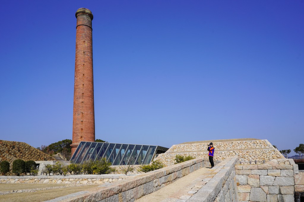 A photo of a low, contemporary granite-stone and glass structure with a tall, much-older smokestake emerging from it.