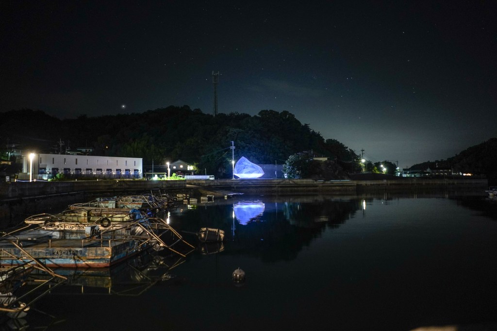A photo of a rural harbor at night. A large geometric form glows with blue light in the distance.
