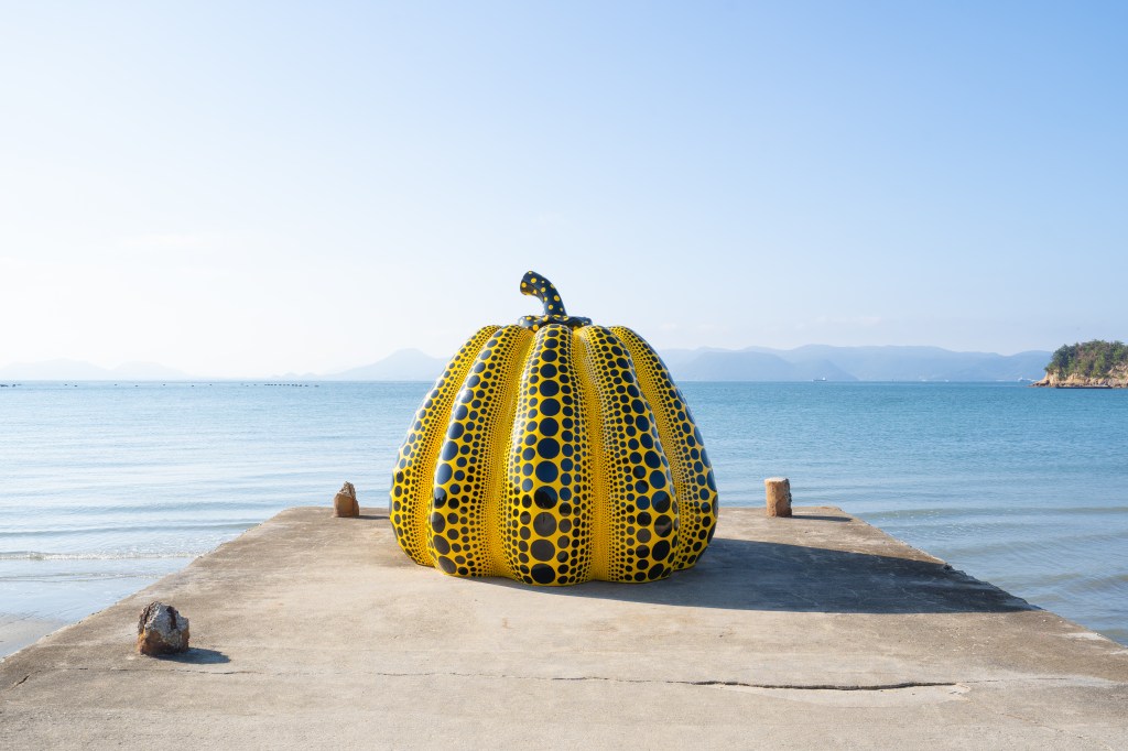 A photo of a large yellow pumpkin sculpture on the end of a pier with the sea in the background.