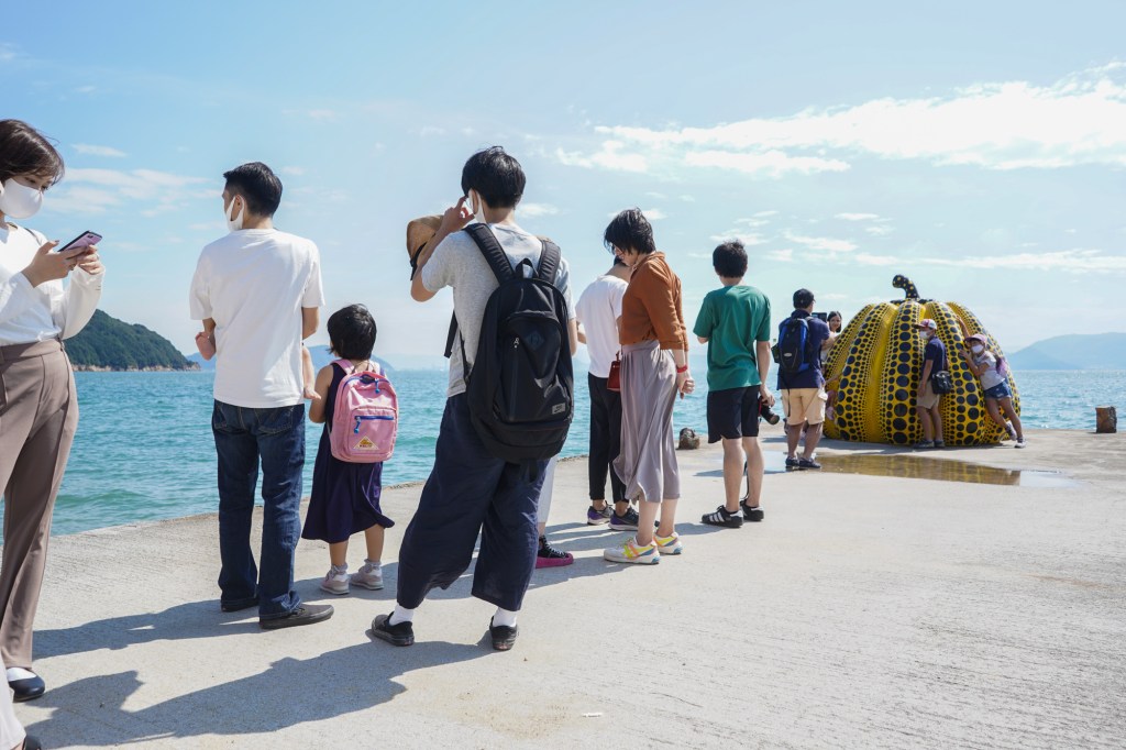 A photo of a line of people standing on a pier, with a yellow pumpkin sculpture in the background.