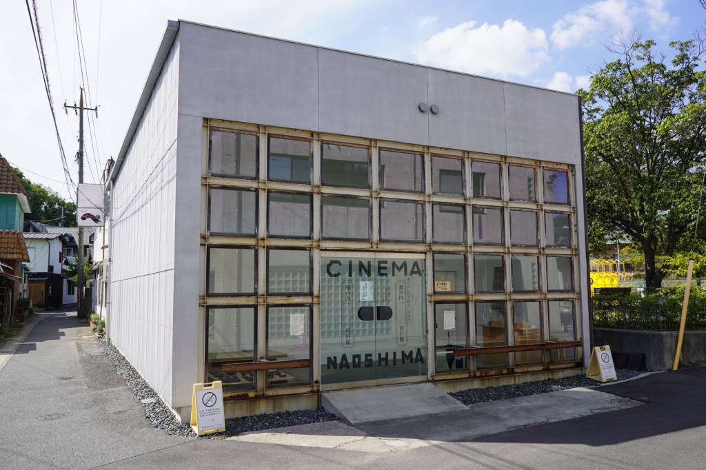 A photograph of a cube-shaped building with a grid of square windows on the front. The glass doors are adorned with large black letters that say "CINEMA NAOSHIMA."