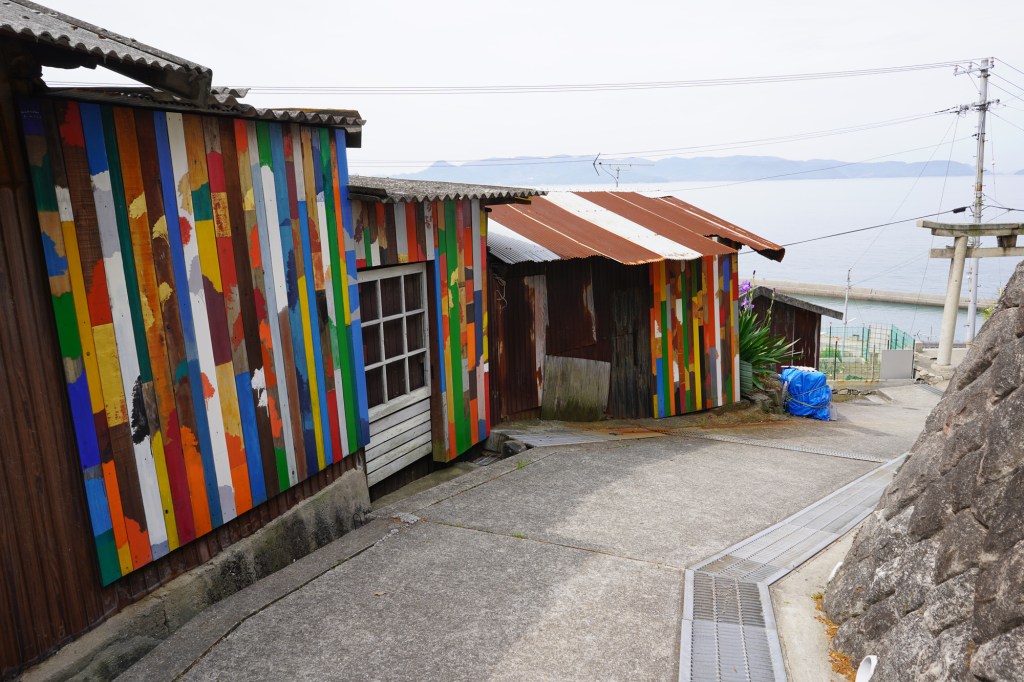 A photo of several small, rustic buildings covered with colorfully-painted vertical boards, with the sea in the background.