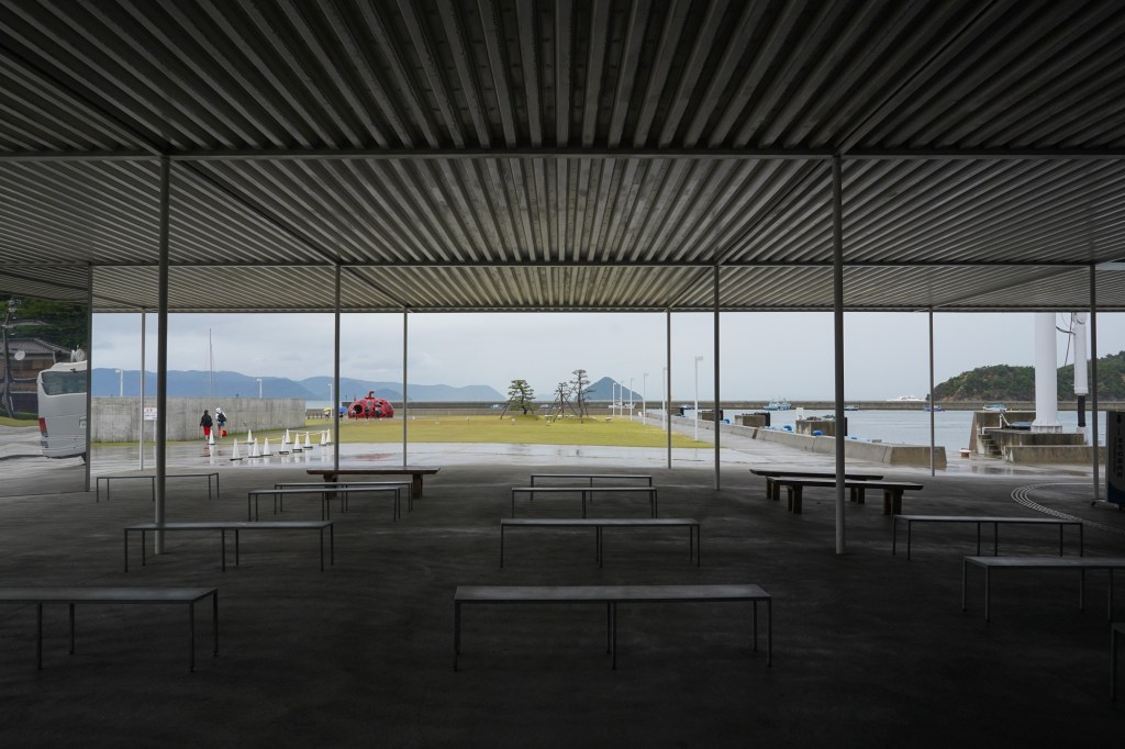 A photo from within a large corrugated-steel shelter, with metal benches on the ground. The sea is visible in the distance.
